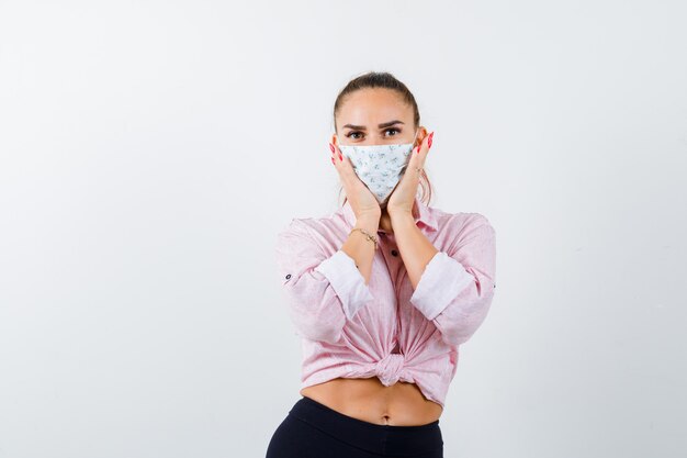 Jeune femme tenant les mains sur les joues en chemise, pantalon, masque médical et à la confiance. vue de face.