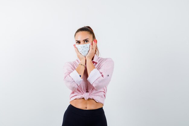 Jeune Femme Tenant Les Mains Sur Les Joues En Chemise, Pantalon, Masque Médical Et à La Perplexité. Vue De Face.