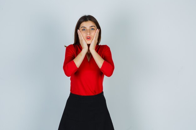 Jeune femme tenant les mains sur les joues en chemisier rouge, jupe et à la perplexité