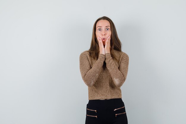 Jeune femme tenant les mains sur le menton en pull doré et pantalon noir et à la surprise