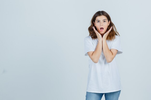 Jeune femme tenant les mains près des joues en t-shirt, jeans et à la surprise. vue de face.