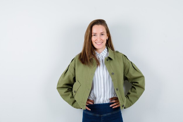 Jeune femme tenant les mains sur la taille en chemise, veste et semblant joyeuse. vue de face.