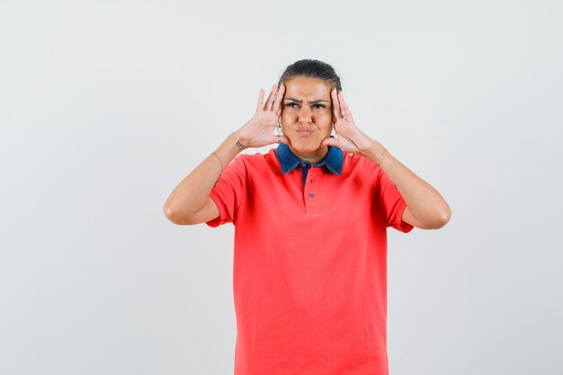 Jeune femme tenant les mains sur la tête, grimaçant en t-shirt rouge et l'air lugubre. vue de face.