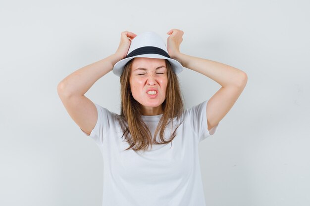 Jeune femme tenant les mains sur la tête en t-shirt blanc, chapeau et à la nostalgie.