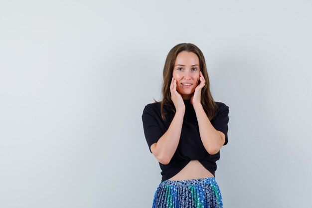 Jeune femme tenant les mains sur le visage, souriant gracieusement en t-shirt noir et jupe bleue et à la recherche attrayante