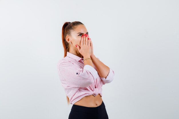 Jeune femme tenant les mains sur le visage tout en regardant en chemise décontractée, pantalon et à la vue étonnée, de face.