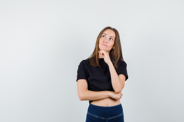 Jeune femme tenant le menton sur place en chemisier noir, pantalon et à la rêveuse