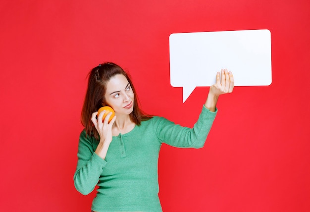 Jeune femme tenant une orange fraîche et un panneau d'information rectangle et a l'air réfléchie