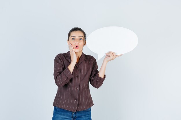 Jeune femme tenant la paume sur la joue, gardant une affiche en papier dans une chemise, un jean et l'air surpris, vue de face.