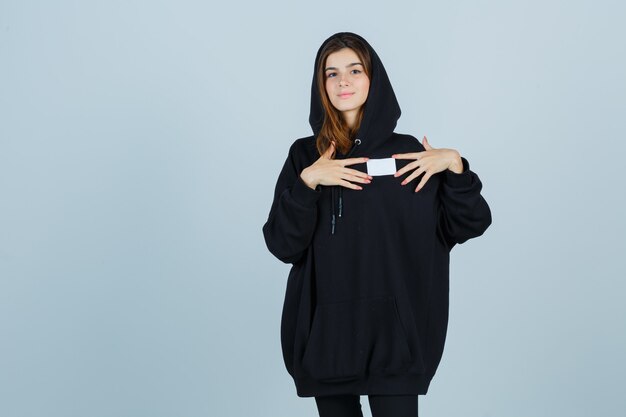 Jeune femme tenant un pense-bête sur la poitrine en sweat à capuche surdimensionné, pantalon et à la confiance. vue de face.