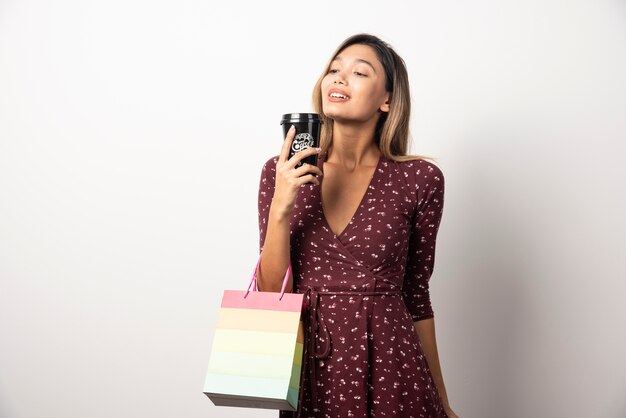 Jeune femme tenant un petit sac de magasin et une tasse de boisson sur un mur blanc.