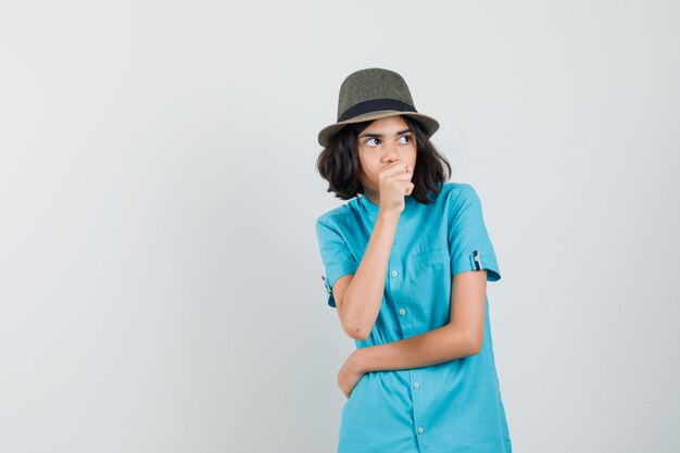 Jeune femme tenant le poing sur sa bouche tout en regardant ailleurs en chemise bleue, chapeau et à la recherche anxieuse.
