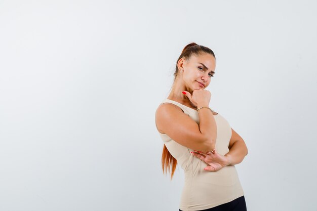 Jeune femme tenant le poing sous le menton en débardeur beige et à la vue de face, confiant.