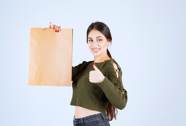 Photo gratuite jeune femme tenant un sac en papier et donnant des pouces sur fond blanc.