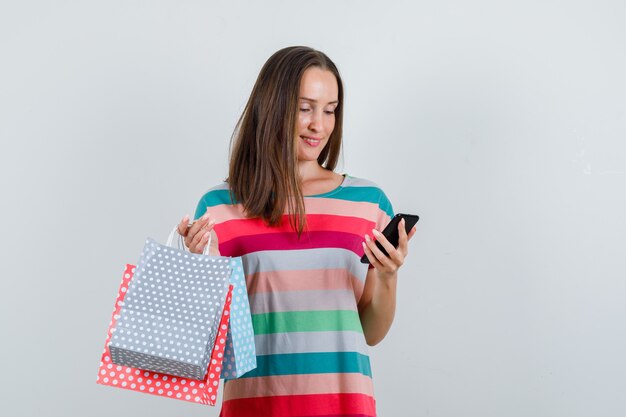 Jeune femme tenant des sacs en papier et regardant le téléphone en vue de face de t-shirt.