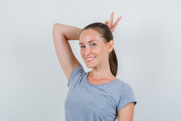 Jeune femme tenant un signe v derrière la tête comme des cornes en t-shirt gris et à la drôle, vue de face.
