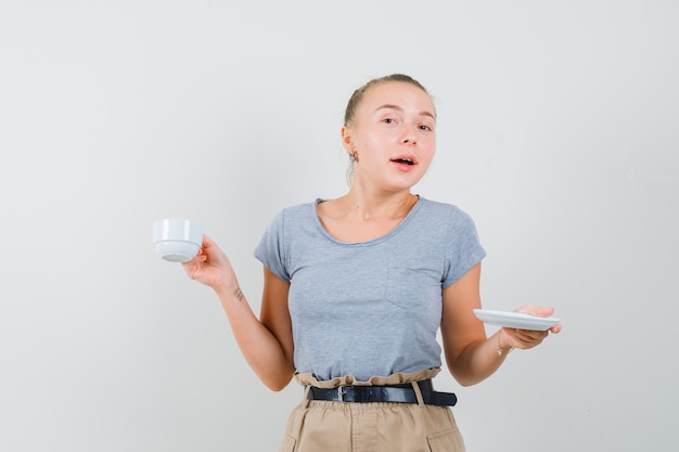 Jeune femme tenant une tasse de boisson en t-shirt, pantalon et à la confusion, vue de face.