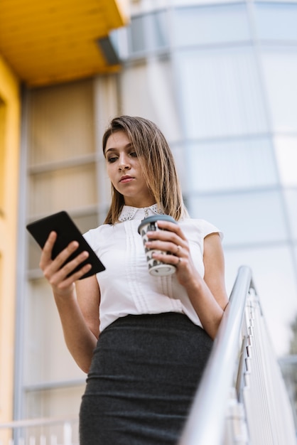 Jeune femme tenant une tasse de café jetable en regardant une tablette numérique