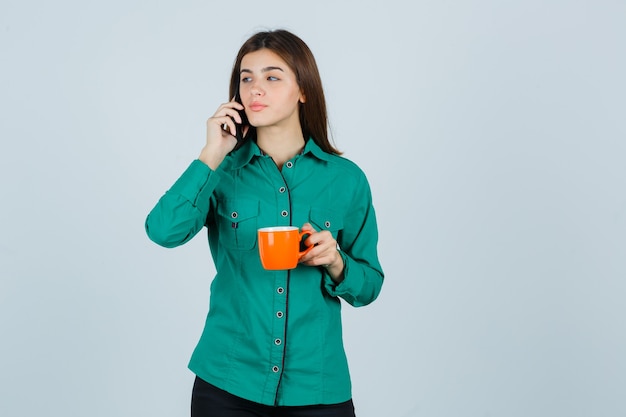 Jeune femme tenant une tasse de thé orange, parlant au téléphone mobile en chemise et à la confiance. vue de face.