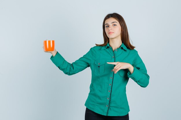 Jeune femme tenant une tasse de thé orange, pointant vers le côté gauche en chemise et à la confiance. vue de face.