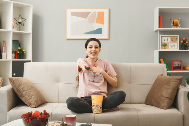 Jeune femme tenant la télécommande de la télévision à la caméra assise sur un canapé derrière une table basse dans le salon