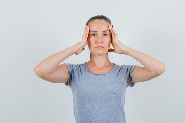 Jeune femme tenant la tête avec les mains en t-shirt gris et à la vue fatiguée, de face.