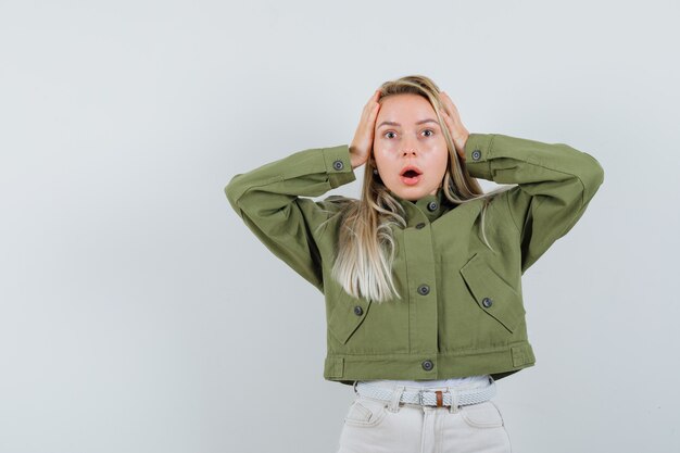 Jeune femme tenant la tête avec les mains en veste verte, jeans et à la vue stressante, de face.