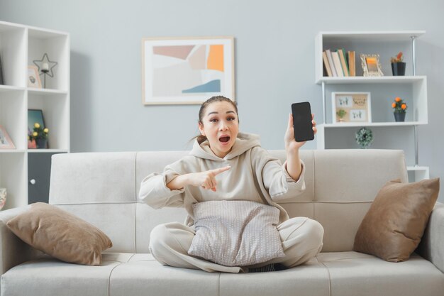 Jeune femme en tenue décontractée assise sur un canapé à l'intérieur de la maison montrant un smartphone pointant avec l'index sur elle étant étonnée et surprise