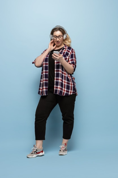 Jeune femme en tenue décontractée avec smartphone et casque sur mur bleu. Caractère positif du corps, féminisme, s'aimer, concept de beauté. Taille plus belle femme d'affaires. Inclusion, diversité.