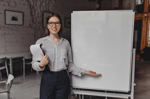 Jeune femme en tenue noir et blanc et lunettes avec sourire regarde dans la caméra détient des documents et pointe vers le tableau de bureau blanc