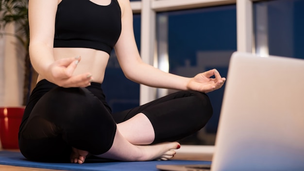 Jeune femme en tenue de sport médite sur un tapis de yoga avec ordinateur portable en face d'elle