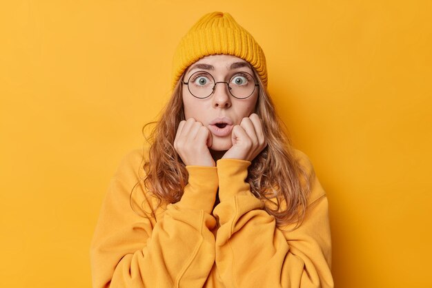 Une jeune femme terrifiée et choquée garde la bouche ouverte vérifie quelque chose d'horrible fixe les yeux sur écoute tient les mains sur les joues porte un pull chapeau et des lunettes rondes isolées sur fond jaune