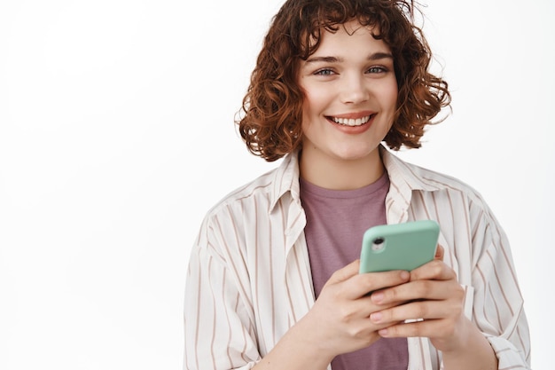 Jeune femme textos sur téléphone mobile. Une fille sourit à la caméra, utilise un smartphone, lit un message ou utilise une application, commande une application de taxi, debout sur fond blanc