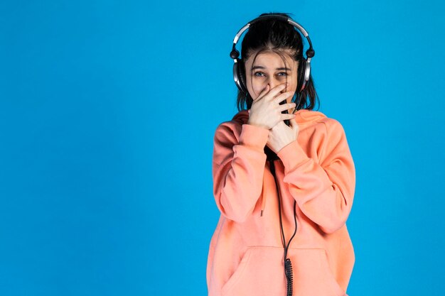 Jeune femme timide portant des écouteurs et mettant ses mains sur sa bouche sur fond bleu Photo de haute qualité