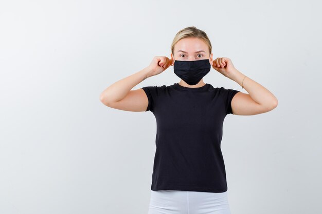 Jeune femme tirant les lobes des oreilles avec les doigts en t-shirt, pantalon, masque médical et ayant l'air drôle