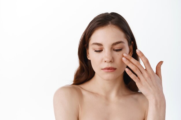 Jeune femme touchant doucement la peau douce et hydratée du visage, appliquant une crème hydratante ou des cosmétiques de soin de la peau, portant un maquillage naturel, debout avec les épaules nues, mur blanc