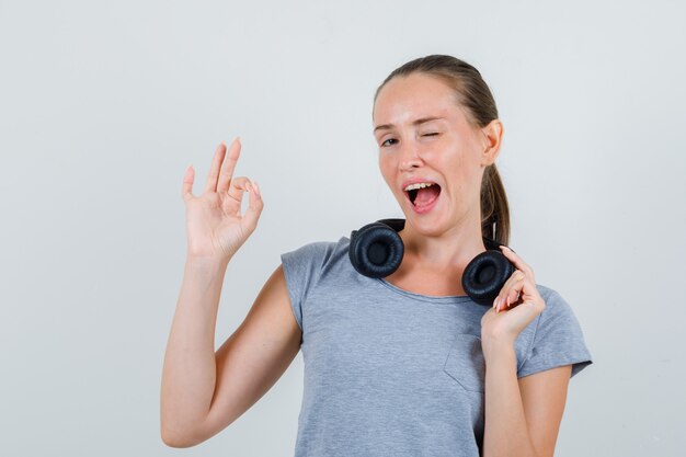 Jeune femme touchant les écouteurs avec un geste ok en vue de face de t-shirt gris.