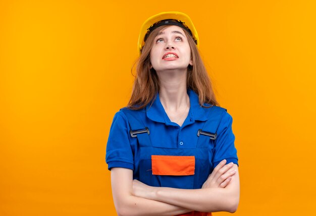 Jeune femme travailleur constructeur en uniforme de construction et casque de sécurité debout avec les bras croisés à la recherche sur le mur orange