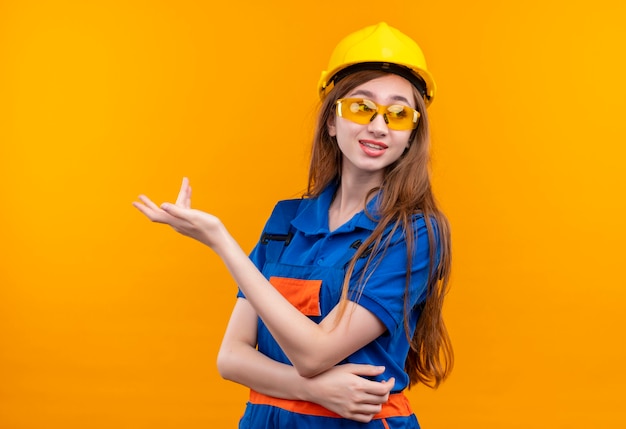 Jeune femme travailleur constructeur en uniforme de construction et casque de sécurité debout avec la main levée en faisant valoir avec une expression sceptique debout sur un mur orange