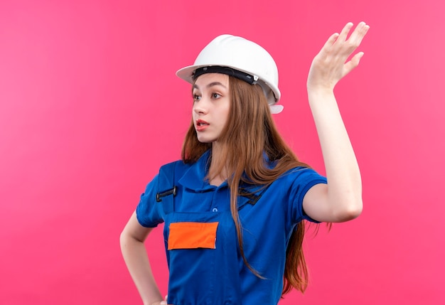 Jeune femme travailleur constructeur en uniforme de construction et casque de sécurité à la main levée confus et mécontent debout sur le mur rose