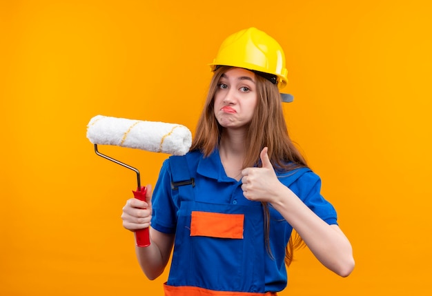 Jeune femme travailleur constructeur en uniforme de construction et casque de sécurité tenant le rouleau à peinture montrant les pouces vers le haut debout sur le mur orange