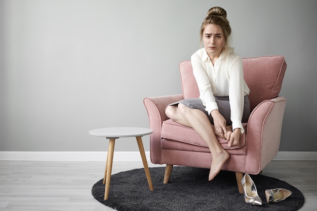 Photo gratuite jeune femme travailleuse moderne se sentant fatiguée après une longue marche sur des chaussures à talons hauts, assise pieds nus dans un fauteuil, massant ses pieds, ayant une expression faciale douloureuse et frustrée. santé et bien-être