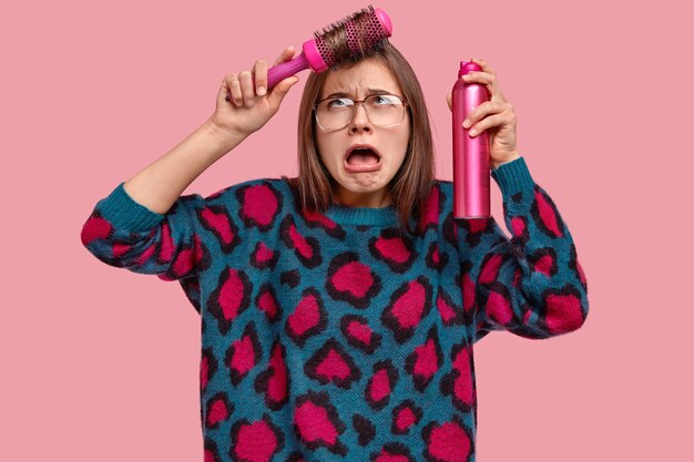 Une jeune femme triste a les cheveux foncés, en colère car elle ne peut pas faire une bonne coiffure, tient de la laque, porte un pull à pois, mécontentement de la qualité des cheveux