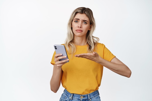 Jeune femme troublée demandant de l'aide avec un téléphone portable pointant vers un smartphone et regardant bouleversée debout sur fond blanc