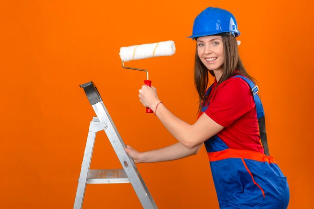 Jeune femme en uniforme de construction et casque de sécurité bleu sur l'échelle en souriant et tenant le rouleau à peinture sur fond orange