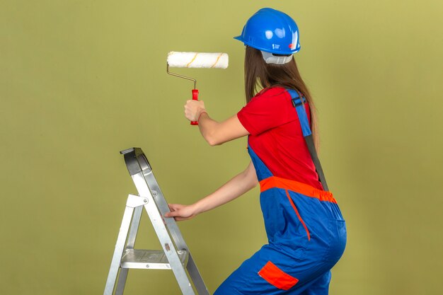 Jeune femme en uniforme de construction et casque de sécurité bleu sur l'échelle tenant le rouleau à peinture sur fond vert