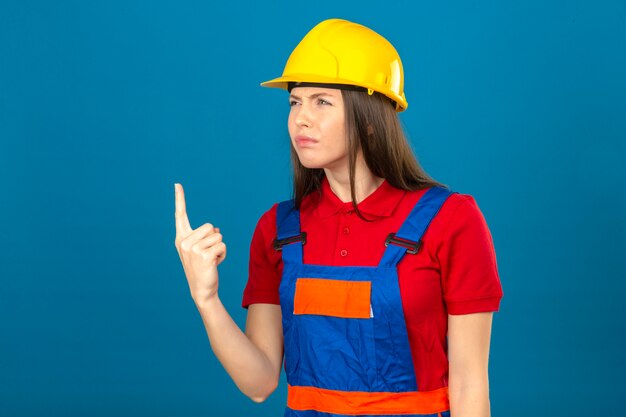 Jeune femme en uniforme de construction et casque de sécurité jaune montrant la déception du doigt pointé vers le haut malheureux à la recherche debout sur fond bleu