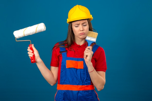 Jeune femme en uniforme de construction et casque de sécurité jaune pensant avec expression pensive visage sérieux tenant un rouleau à peinture et un pinceau debout sur fond bleu