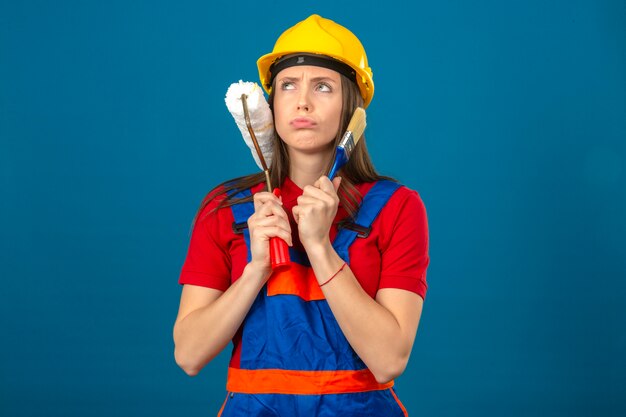 Jeune femme en uniforme de construction et casque de sécurité jaune pensant avec expression pensive visage sérieux tenant un rouleau à peinture et un pinceau debout sur fond bleu