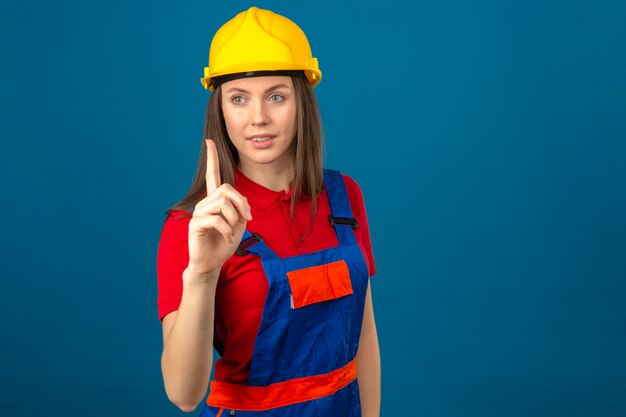 Jeune femme en uniforme de construction et casque de sécurité jaune pointant vers le haut avec le doigt souriant debout sur fond bleu